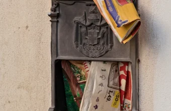 An ornate cast-iron wall mailbox with its door hanging open, stuffed with colorful flyers and a yellow flyer bulging out of the top slot