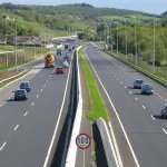 A dual carriageway seen from above, with a grass and concrete-barrier median strip separating the two carriageways and a 100 km/h speed limit sign standing on the median
