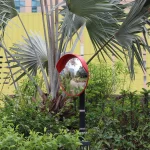 Red convex traffic mirror on a post among palm fronds and shrubs, showing a small curved reflection of a garden path
