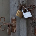 Two padlocks with interlocked shackles, one brass and one steel, securing the weathered door of an old gate