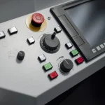 Close-up of an industrial machine operator control panel with a red emergency stop button, push buttons, a handwheel and a display screen