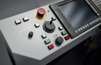 Close-up of an industrial machine operator control panel with a red emergency stop button, push buttons, a handwheel and a display screen
