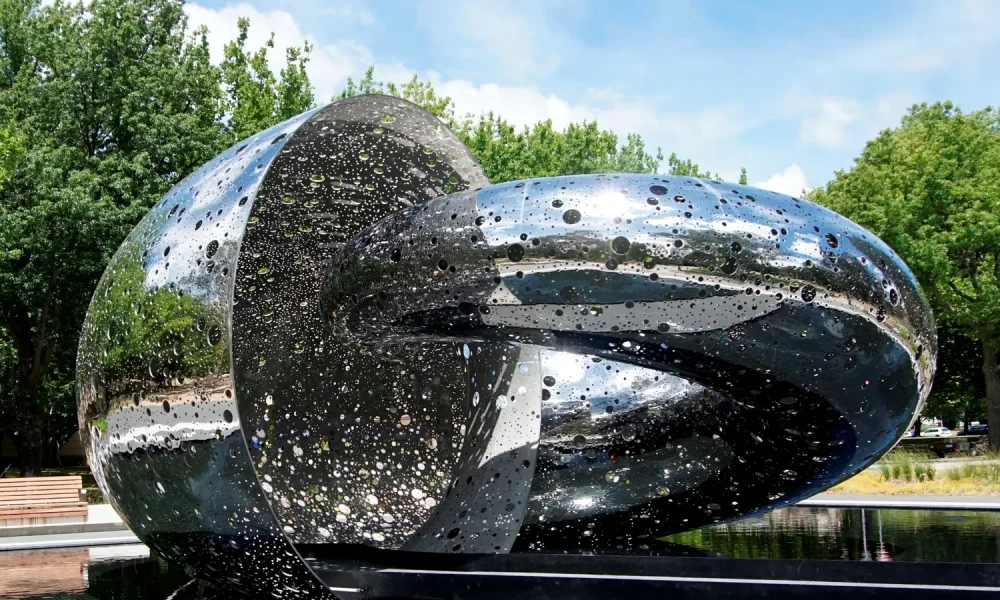 Ouroboros, a polished stainless steel sculpture of two interlocking loops with a pierced surface, standing beside a dark pool under trees