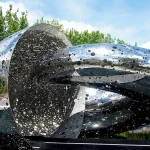Ouroboros, a polished stainless steel sculpture of two interlocking loops with a pierced surface, standing beside a dark pool under trees