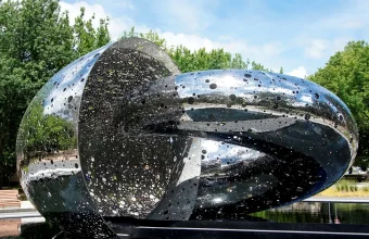 Ouroboros, a polished stainless steel sculpture of two interlocking loops with a pierced surface, standing beside a dark pool under trees