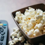 A TV remote control beside a bowl of popcorn on a dark wooden table