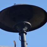 A black public warning siren with a wide dish-shaped horn on a short pole against a clear blue sky