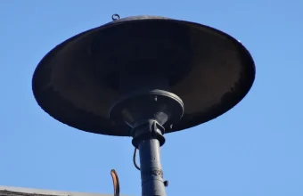 A black public warning siren with a wide dish-shaped horn on a short pole against a clear blue sky