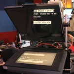 A broadcast studio teleprompter with script lines shown mirrored on its lower monitor and reflected in the glass in front of a camera lens