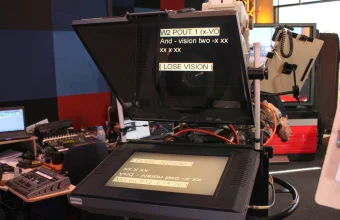 A broadcast studio teleprompter with script lines shown mirrored on its lower monitor and reflected in the glass in front of a camera lens