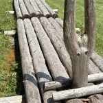 A raft of long grey wooden logs lying in a green meadow, with a forked post standing in the middle