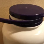A purple bottle cap with its tamper-evident ring torn free and hanging off to one side, photographed against a beige wall