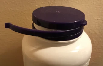 A purple bottle cap with its tamper-evident ring torn free and hanging off to one side, photographed against a beige wall
