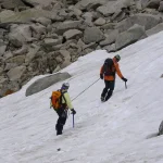 Two climbers in helmets and backpacks walk up a snow slope, joined by a taut rope between them