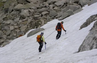 Two climbers in helmets and backpacks walk up a snow slope, joined by a taut rope between them