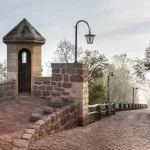 A stone sentry box on a castle wall beside a lamp post and a cobblestone path in morning mist at Wartburg Castle