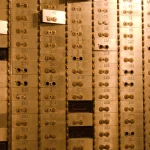 A brass wall of numbered bank safe deposit boxes, each with a pair of keyholes