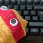 A red and yellow striped sock puppet with googly eyes resting on a black computer keyboard