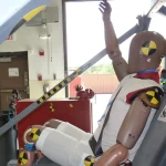 A crash test dummy with one arm raised, seated in a test rig after a simulated frontal crash, with yellow and black markers on its head, shoulder and knees