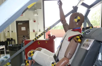 A crash test dummy with one arm raised, seated in a test rig after a simulated frontal crash, with yellow and black markers on its head, shoulder and knees