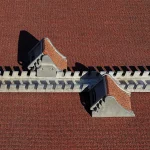Overhead view of an athletics starting block, with two angled foot plates on a slotted metal rail, lying on a red running track