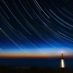 Long-exposure photo of curved star trails above a lighthouse and the sea at dusk