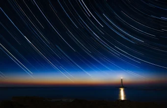 Long-exposure photo of curved star trails above a lighthouse and the sea at dusk