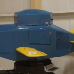 A blue Link Trainer flight simulator with a yellow wing and tail on a pedestal in a museum hangar