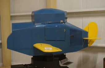 A blue Link Trainer flight simulator with a yellow wing and tail on a pedestal in a museum hangar