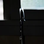 A metal tap in silhouette beside a window with drops of water falling from its spout