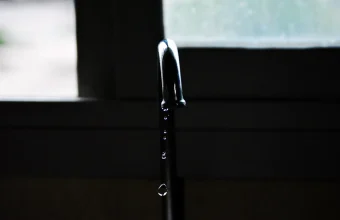 A metal tap in silhouette beside a window with drops of water falling from its spout