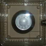 A round wall thermostat behind a clear locked plastic cover, its temperature dial visible but out of reach