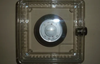 A round wall thermostat behind a clear locked plastic cover, its temperature dial visible but out of reach