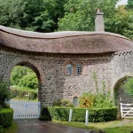 A stone gatehouse with a thatched roof and a white toll gate across a road in Somerset, England