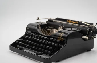 A black 1935 Olympia Simplex portable typewriter with gold lettering, photographed against a plain light grey background
