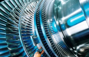 A worker installs a blade on a large steam turbine rotor in a factory, with rings of polished blades curving across the frame