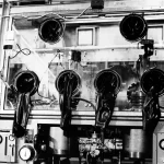 Black rubber gloves hanging through round ports in the glass front of a large laboratory glove box, shown in a 1966 black-and-white photograph