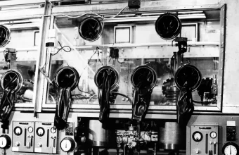 Black rubber gloves hanging through round ports in the glass front of a large laboratory glove box, shown in a 1966 black-and-white photograph