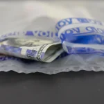 Clear tamper-evident security bag with blue VOID tape peeled back and a folded US hundred-dollar bill inside