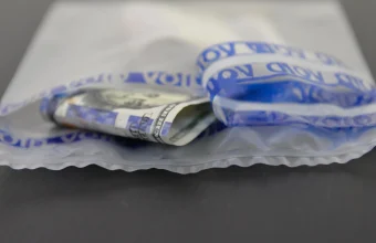 Clear tamper-evident security bag with blue VOID tape peeled back and a folded US hundred-dollar bill inside