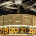 Close view of the Urania World Clock in Berlin at night, with city names grouped by time zone above a band of gold hour numerals from 17 to 24